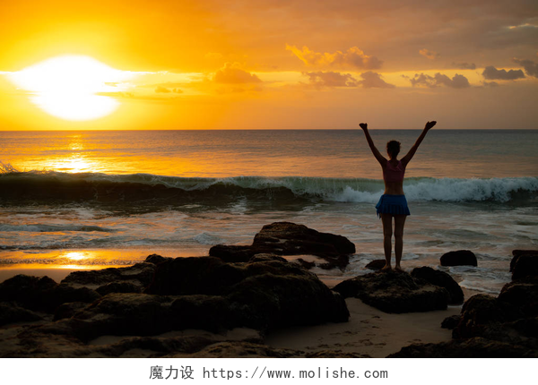 黄昏下年轻女子站在海边的岩石上兴奋的年轻女子在海边的海滩上举起双臂。从后面查看。日落在海滩。巴厘岛, 印度尼西亚.